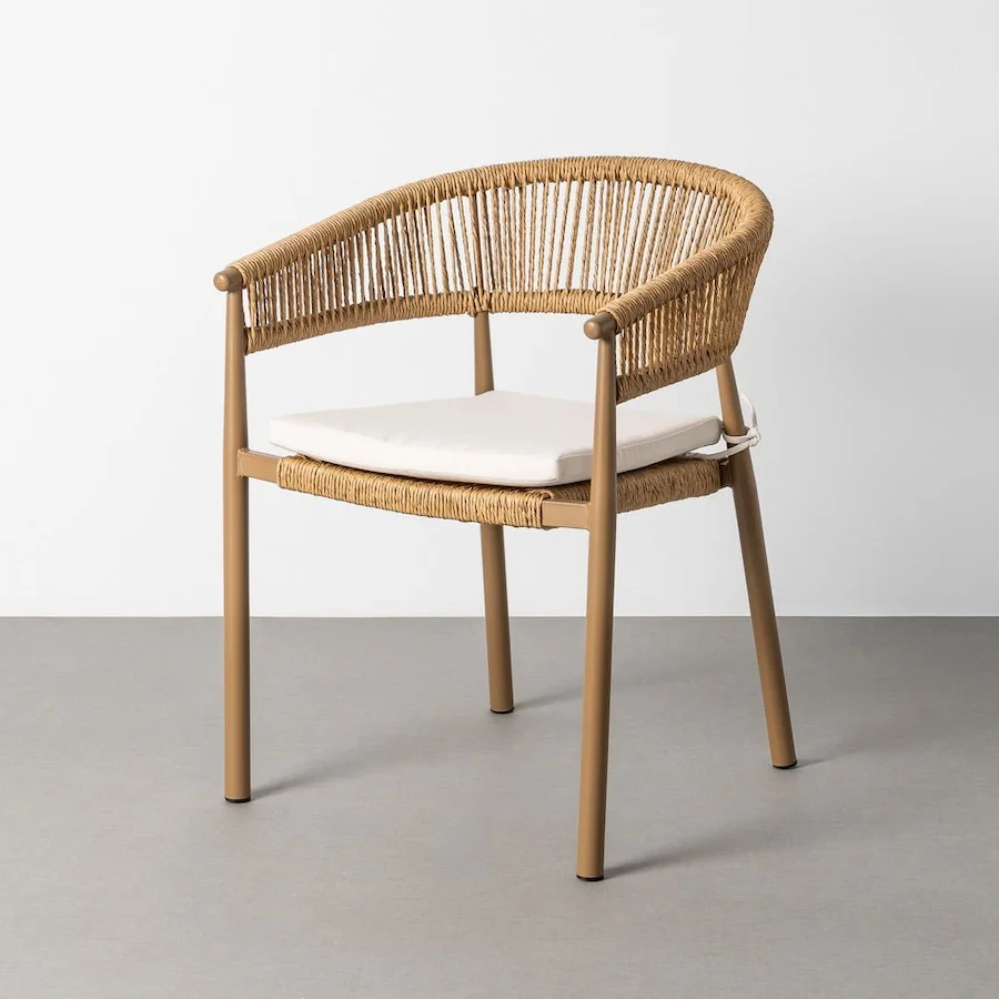 Chaise de jardin à dossier arrondi en rotin synthétique, avec accoudoirs et coussin d'assise clair, devant un mur blanc